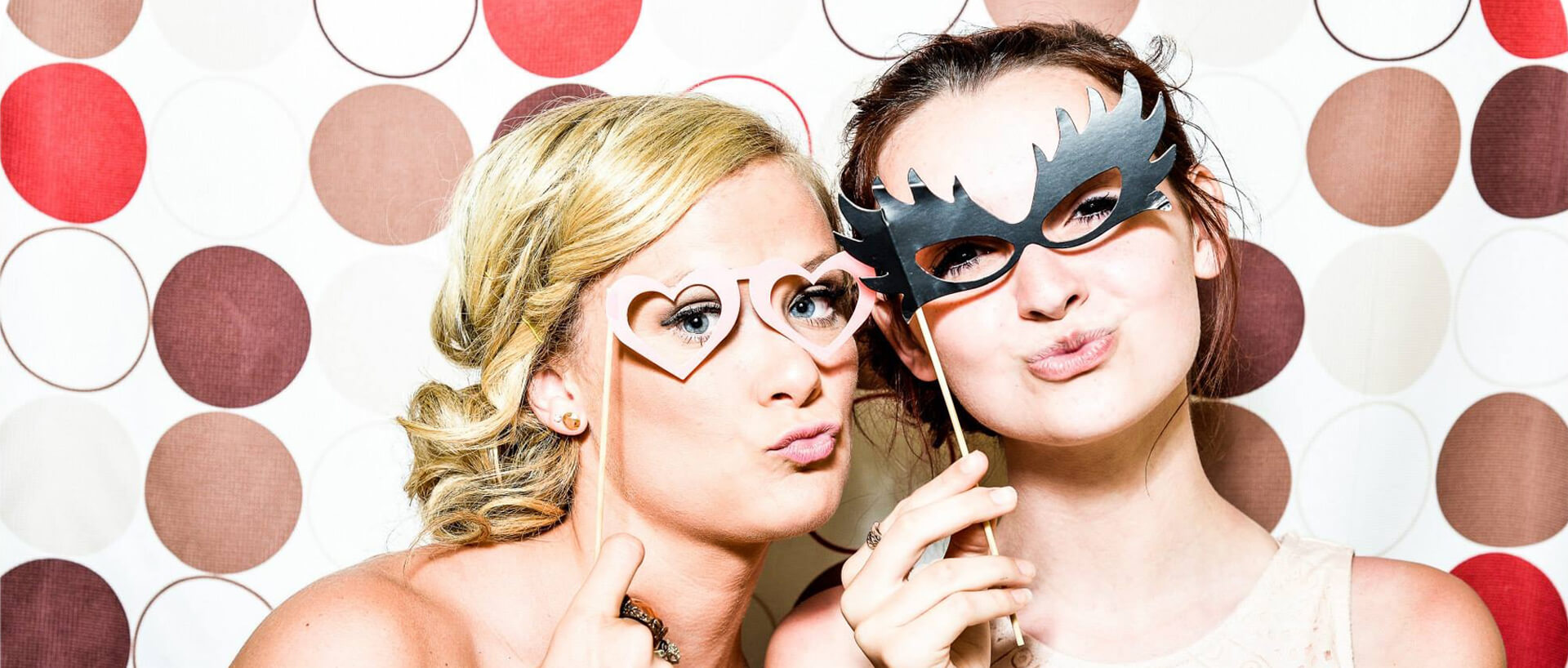 Two women make fun faces with props against a wall of a photo-booth.