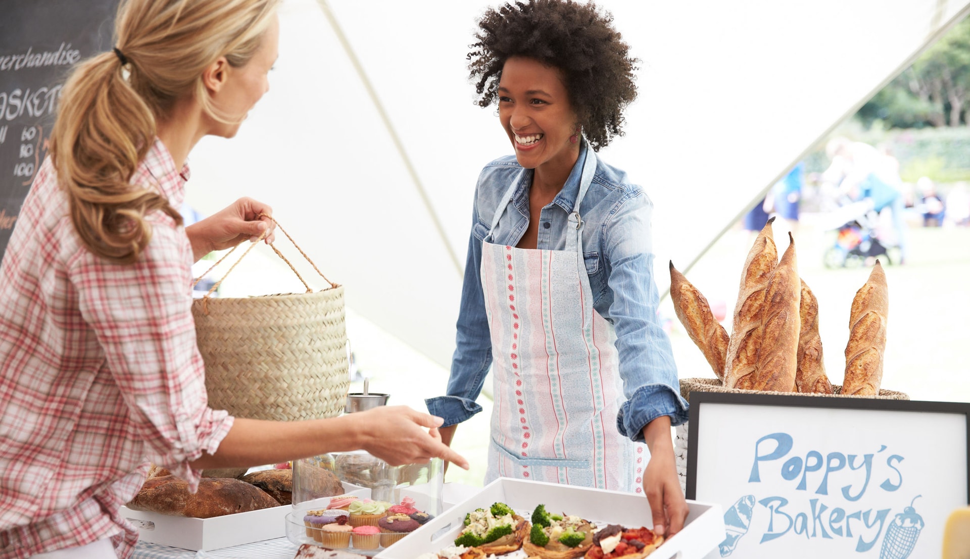 A woman business owner at a bakery stand smiles and offers a sample to a customer.