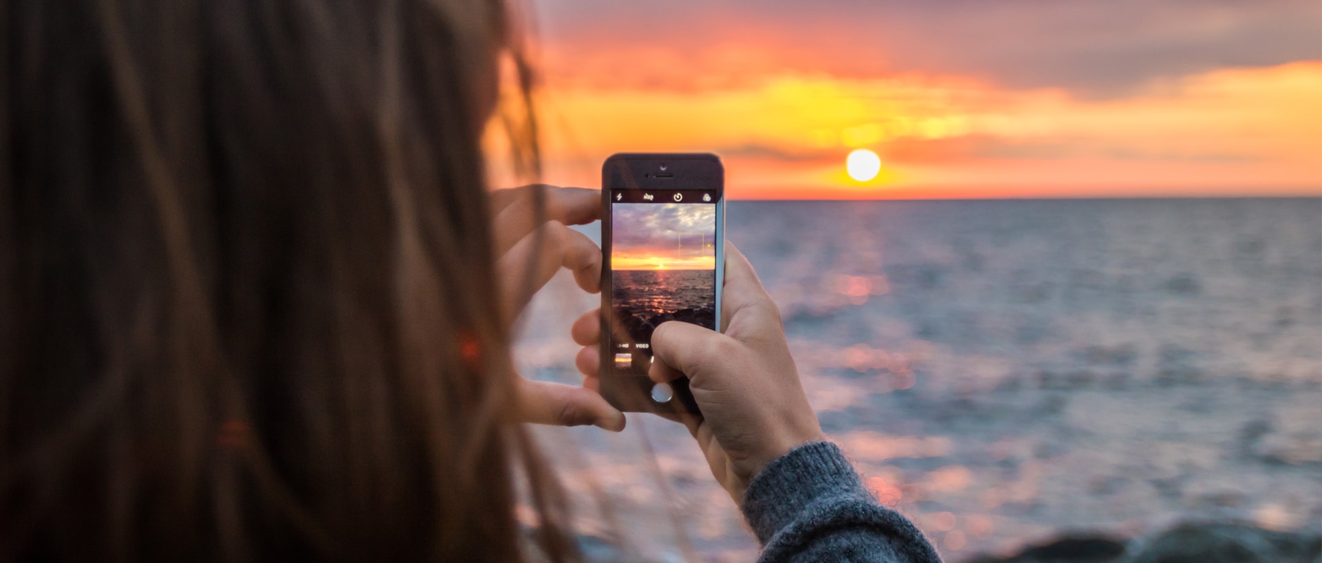 A woman holding up her phone to take a photo of a sunset.